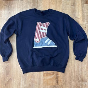 May include: A navy blue crewneck sweatshirt featuring a large, centered fabric appliqué of a high-top sneaker. The sneaker design is composed of red and white vertical stripes on the upper section and a solid light blue lower section.