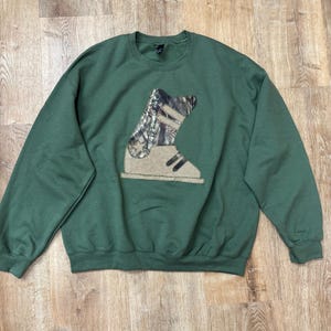 May include: A forest green long-sleeve sweatshirt featuring a large central applique of a ski boot. The boot design is split into two sections: a camouflage patterned upper portion and a solid tan lower portion with dark strap details.