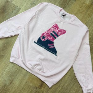 May include: A light pink crewneck sweatshirt featuring a large central graphic of a boot. The boot design combines a dark blue denim-textured lower section with a bright pink and white paisley bandana-patterned upper section and a star detail.