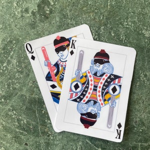 May include: Two playing cards, a Queen and a King, rest on a green surface. The cards feature illustrations of figures wearing sunglasses and holding objects. The Queen holds a pink object, and the King holds a gray object.