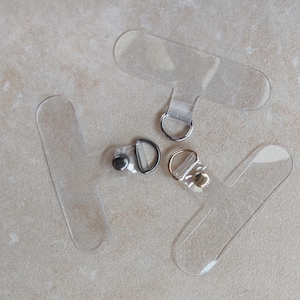 Könnte beinhalten: Vier transparente Plastikbänder mit Metall- und goldfarbenen Verschlüssen. Die Bänder sind dazu bestimmt, Gegenstände an einer Oberfläche zu befestigen.