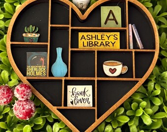 May include: A heart-shaped wooden shelf with multiple compartments, painted black inside. It contains miniature items like a cactus, a blue vase, a book, and a coffee cup. The shelf has the text "ASHLEY'S LIBRARY" and "book lover".