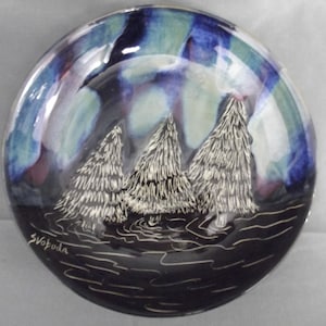 May include: A ceramic plate with a winter scene. The plate features three white fir trees against a backdrop of blue, green, and purple hues. The plate's rim is a darker blue, and the artist's signature is visible.
