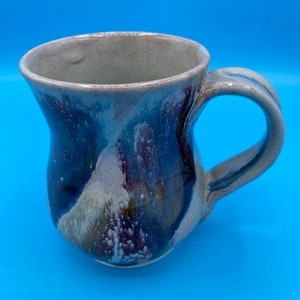 May include: A ceramic mug with a flared base and a handle. The mug has a light gray interior and a blue, purple, and white glaze on the exterior.