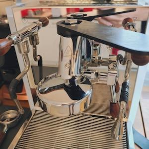 Può includere: Macchina per caffè espresso cromata e in legno con un vassoio raccogli gocce in acciaio inossidabile. La macchina ha un manometro e una lancia vapore. La macchina è su una superficie bianca.