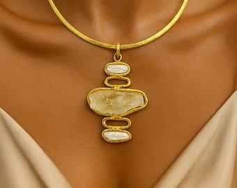 May include: A gold-toned necklace with a pendant featuring a large, irregularly shaped, light yellow stone, and two smaller white oval stones. The necklace has a hammered texture and is displayed on a person.