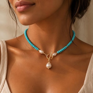 Op de afbeelding: Een ketting bestaande uit een reeks kleine, ronde, fel turquoise kralen, geaccentueerd met witte, onregelmatig gevormde parels. Het sieraad is voorzien van een goudkleurige toggle-sluiting in het midden met een enkele hangende parel.