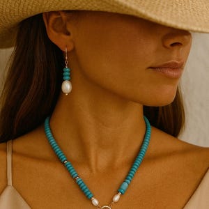 Puede incluir: Un collar de cuentas turquesas y pendientes a juego con detalles de perlas. El collar presenta un cierre dorado y un gran colgante de perla. Los pendientes tienen un gancho dorado y una gota de perla. La joya se lleva con un sombrero de paja.