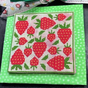 May include: A green and white cross-stitch piece with a design of red strawberries on a white background. The piece is framed in a black frame and is sitting on a green surface.
