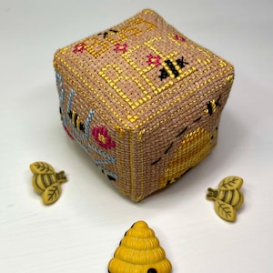 Puede incluir: Un cubo de punto de cruz beige con diseños de abejas y panales amarillos y negros. Dos botones con forma de abeja y un botón con forma de colmena amarilla están dispuestos alrededor del cubo. El cubo mide aproximadamente 7.6 cm de ancho.