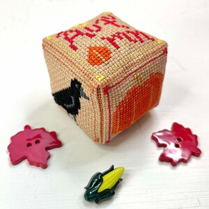 Op de afbeelding: Een klein, vierkant, beige kussen met een kruissteekontwerp met de woorden "Happy Fall Y'all" in rood en oranje garen. Het kussen is omringd door drie rode esdoornbladknoppen en een groene maïs knop.