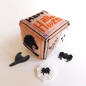 Op de afbeelding: Een beige kubus met de woorden "Happy Halloween" erop geborduurd. De kubus wordt omringd door een zwarte heksenhoed, een wit spinnenweb met een zwarte spin en twee zwarte spinnen.