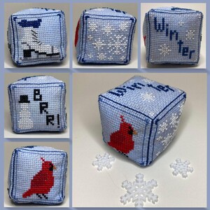 May include: A light blue fabric cube featuring cross-stitch embroidery of winter themes including a white ice skate, white snowflakes, a snowman with the text BRR, and a red cardinal bird. The word Winter is stitched in dark blue on the top face.