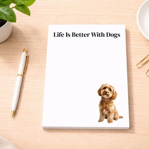 May include: A white notepad with the text "Life Is Better With Dogs" and a brown dog illustration. A white pen with gold accents and a small potted plant are on the left. Gold paper clips and a cup of coffee are on the right.