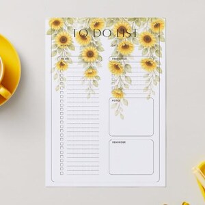 Op de afbeelding: Een witte To Do List-notitieblok met een zonnebloemontwerp. Het notitieblok heeft secties voor maand, datum, prioriteiten, notities en herinneringen. Een gele kop en schotel bevinden zich in de linkerbovenhoek.