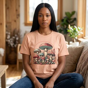Op de afbeelding: Een perzikkleurig T-shirt met korte mouwen en een centrale grafische illustratie van diverse paddenstoelen in rood-, blauw- en crèmetinten, gerangschikt onder een halfrond boogontwerp met gedetailleerde botanische elementen.
