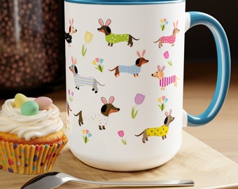 May include: White ceramic mug with a blue rim and handle. The mug features a repeating pattern of dachshunds wearing bunny ears and colorful pajamas. The dachshunds are surrounded by flowers and Easter eggs.
