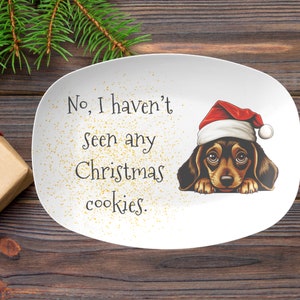 May include: White ceramic serving dish with a brown and white dog wearing a red Santa hat. The dish has gold glitter and the text "No, I haven't seen any Christmas cookies."