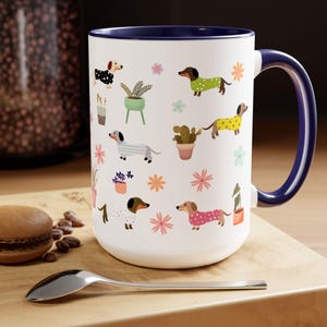 May include: White ceramic coffee mug with a blue rim and handle. The mug features a repeating pattern of dachshunds wearing colorful clothes and potted plants.