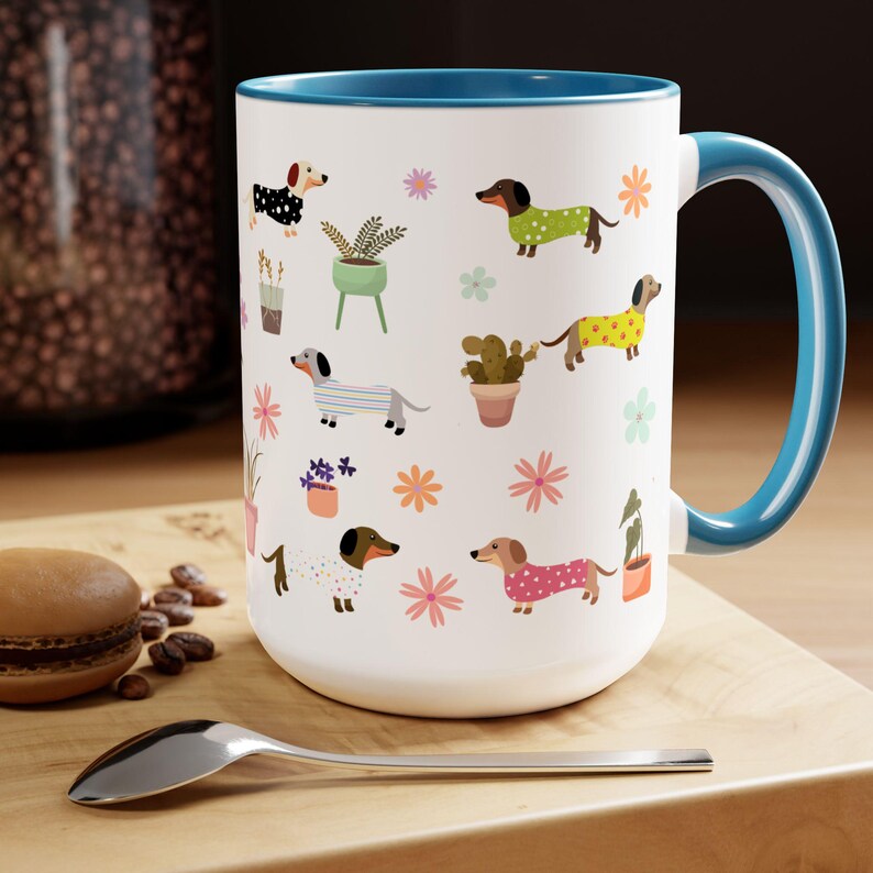 May include: White ceramic coffee mug with a blue rim and handle. The mug features a repeating pattern of dachshunds wearing colorful clothes and potted plants.