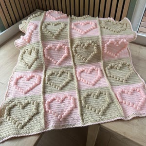 Può includere: Una coperta all'uncinetto fatta a mano con un motivo a cuore. La coperta alterna quadrati rosa e beige, ognuno con un motivo a cuore in rilievo. La coperta è realizzata in filato morbido, adatta a un bambino. Un oggetto decorativo e accogliente.