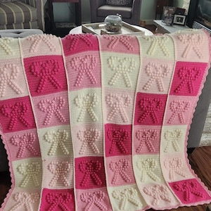 May include: A crocheted baby blanket with a pink and white color scheme. The blanket features a repeating pattern of bows with a textured, puffy look.