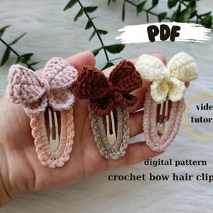 May include: Three crocheted bow hair clips in pink, brown, and white. The clips are attached to a metal bar and are ready to be used. The image includes the text "PDF", "video tutorial", and "digital pattern crochet bow hair clips".