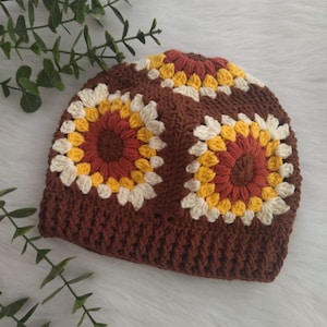 Puede incluir: Un gorro de punto marrón con dos grandes diseños de flores de colores. Las flores están hechas con hilo blanco, amarillo, naranja y marrón.