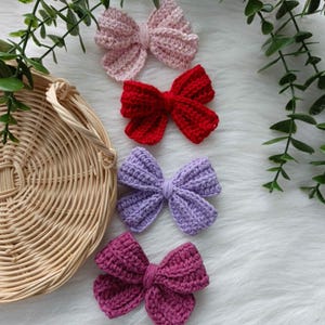 May include: Four crocheted bows in different colours: pink, red, purple, and burgundy. The bows are arranged in a vertical line on a white fluffy surface.