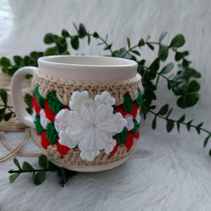 Puede incluir: Una taza de cerámica blanca con un acogedor de ganchillo. El acogedor es beige con rayas rojas, blancas y verdes y una flor blanca de ganchillo.