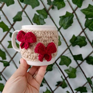 May include: A white ceramic mug with a crocheted beige and red bow design. The mug is being held by a hand.