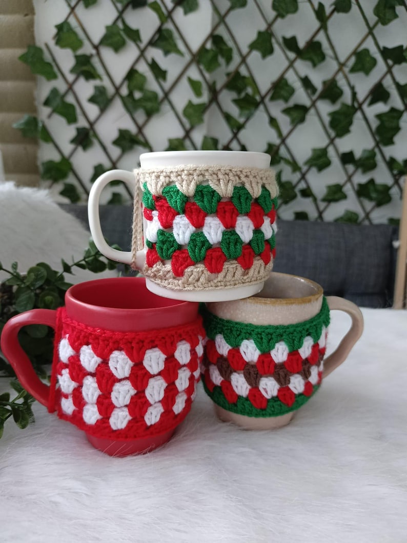May include: Three ceramic mugs with crocheted cozies.  One cozy is beige with green and red diamond pattern, another is red and white, and the third is beige with brown, green, and red diamond pattern. The mugs and cozies are perfect for winter holidays.