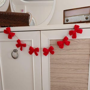 May include: A red knitted bow garland with six bows strung on a twine string. The garland is hanging on a white cabinet with a wood grain panel.