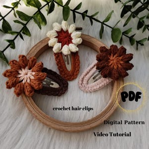 May include: Three crocheted flower hair clips in shades of brown, white and cream. The clips are displayed on a wooden hoop. The text "crochet hair clips", "Digital Pattern", and "Video Tutorial" are visible below the clips.