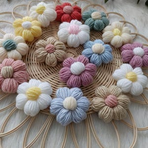 May include: A collection of 14 crocheted flower hair clips in various colors, including pink, blue, yellow, green, and beige. The flowers are arranged on a woven basket.