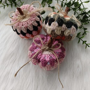 May include: Three crocheted pumpkins in pink, orange, and brown with a rustic twine bow. The pumpkins are on a white fluffy surface.