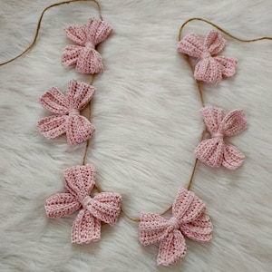 Può includere: Una ghirlanda di fiocchi rosa all'uncinetto infilati su uno spago. I fiocchi sono tutti della stessa dimensione e forma, e sono disposti in uno schema simmetrico.