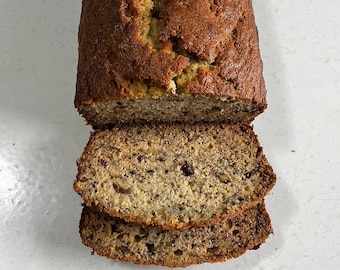 May include: A loaf of freshly baked banana bread, with two slices cut and stacked in front. The bread has a golden-brown crust and a moist interior with visible banana and nut inclusions. The bread sits on a white surface.