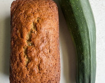 Puede incluir: Un pan dorado junto a un calabacín verde. El pan tiene una superficie texturizada y es rectangular. El calabacín es largo y cilíndrico, de color verde oscuro y con tallo.