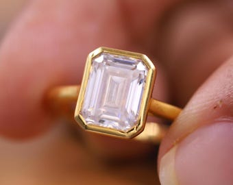May include: A gold ring featuring a large, rectangular, clear gemstone with a gold bezel setting. The ring's band is also gold. The ring is held between two fingers, showcasing the ring's design and details.