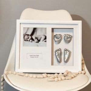 May include: A white frame containing a black and white photograph of a sleeping baby, along with silver hand and foot prints. The frame includes a label that reads "Lincoln Frances Mackin, December 20th, 2022".