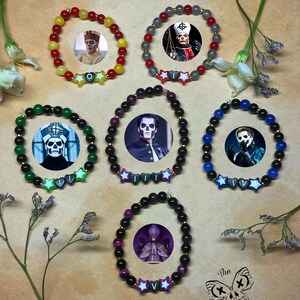 Könnte beinhalten: Eine Sammlung von Armbändern mit Perlen, die Bilder eines Bandmitglieds zeigen. Jedes Armband hat ein anderes Farbschema, darunter rote, gelbe, grüne, schwarze und blaue Perlen. Sternförmige Akzente und das Logo der Band sind ebenfalls zu sehen.
