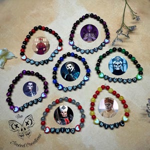 Op de afbeelding: Een verzameling kralenarmbanden in verschillende kleuren, elk met een cirkelvormige afbeelding en een naam die is gespeld met zwarte letterkralen. De armbanden bevatten rode, zwarte, groene en gele kralen, met hartvormige accenten.