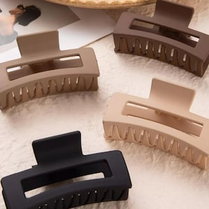 May include: Four rectangular hair claw clips in various neutral colors: black, brown, tan, and beige. Each clip has a matte finish and a series of teeth along the curved edge for securing hair. The clips are arranged on a textured surface.
