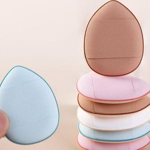 May include: A stack of colorful, teardrop-shaped makeup sponges in various shades of pink, brown, white, and blue. One sponge is held in a hand. The sponges have a textured surface and are designed for applying cosmetics.
