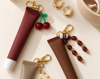 May include: Three squeeze tubes in maroon, brown, and beige, each attached to a gold-tone lobster clasp keychain. The maroon tube features a red cherry charm, the brown tube has a coffee cup charm with brown beads, and the beige tube has a star and pearl charm.