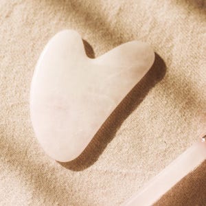 May include: A light pink, heart-shaped gua sha tool made of smooth stone. The tool is designed with curved edges for facial massage and is set against a neutral, textured background. The gua sha is a beauty and wellness product.
