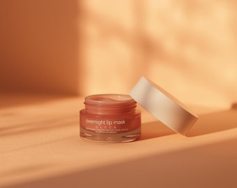 May include: A small, open jar of overnight lip mask with a white lid resting beside it. The jar is filled with a pinkish-red product and has text on the front. The background is a warm, peachy color.