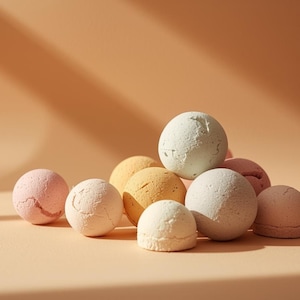 May include: A collection of spherical bath bombs in various pastel colors, including pink, yellow, and white. The bath bombs have a textured surface and are arranged on a light peach background with shadows.