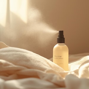 May include: A bottle of pillow spray with a black nozzle is being sprayed, creating a fine mist. The bottle is on a cream-colored blanket. The label reads "Pillow Pillow Spray". The background is a soft, neutral tone.